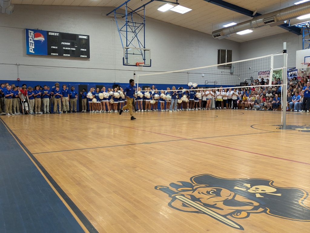 Students and cheerleaders line the walls of a gymnasium while a participant crosses the court during a school assembly.