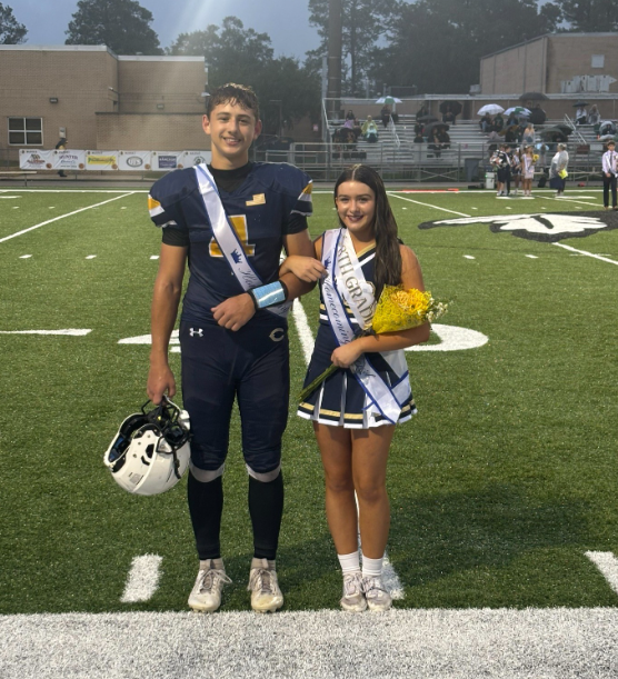 Two middle school homecoming court representatives stand on a football field before a game. One student wears a football uniform with a homecoming sash and holds a football helmet. The other student wears a cheer uniform with a homecoming sash and holds a bouquet of yellow flowers. Bleachers with spectators holding umbrellas are visible in the background, and school buildings surround the field under overcast evening skies.