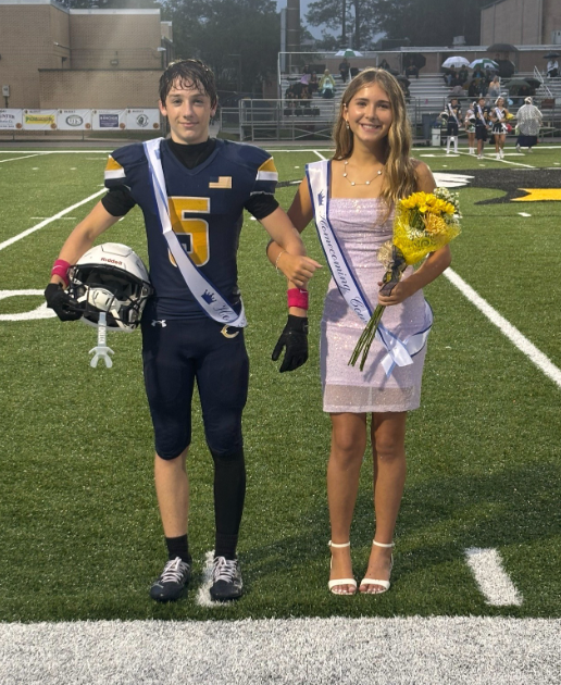 Two middle school homecoming court representatives stand together on a football field before a game. One student wears a football uniform with a homecoming sash and holds a football helmet. The other student wears a light-colored formal dress with a homecoming sash and carries a bouquet of yellow flowers. Spectators holding umbrellas are seated in the bleachers behind them, and school buildings are visible beyond the field under cloudy evening skies.