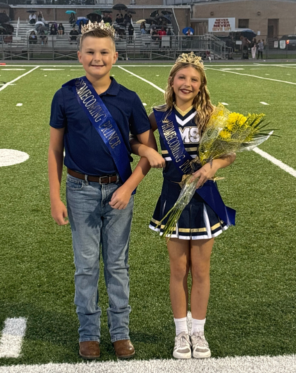 Two middle school homecoming royalty representatives stand together on a football field during an evening ceremony. Both wear crowns and homecoming royalty sashes. One student wears a navy polo shirt, jeans, and boots. The other student wears a cheer uniform and holds a bouquet of yellow flowers. Spectators with umbrellas are seated in the bleachers behind them, and the football field is illuminated by stadium lights.