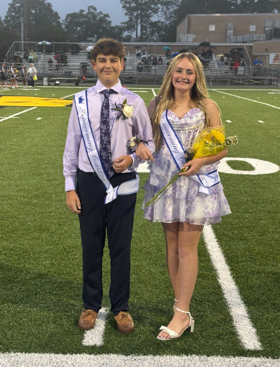 Two middle school homecoming court representatives stand together on a football field during a rainy evening event. Both wear homecoming sashes. One student is dressed in a button-down shirt, tie, dress pants, and a boutonniere. The other student wears a light purple floral dress, white sandals, and carries a bouquet of yellow flowers. Spectators with umbrellas sit in the bleachers behind them, and school buildings are visible beyond the field under overcast evening skies.