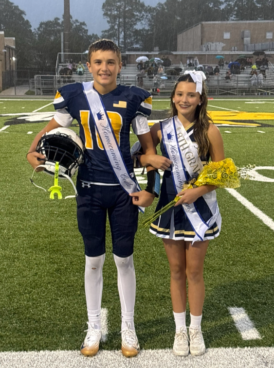 Two middle school homecoming court representatives stand together on a football field during a rainy evening event. One student wears a football uniform with a homecoming sash and holds a football helmet. The other student wears a cheer uniform with a homecoming sash, a large white hair bow, and carries a bouquet of yellow flowers. Rain is visible under the stadium lights, with spectators holding umbrellas in the bleachers behind them.