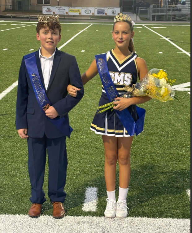 Two middle school homecoming royalty representatives stand together on a football field during an evening ceremony. Both wear crowns and blue homecoming royalty sashes. One student is dressed in a navy suit with a white shirt and brown dress shoes. The other student wears a cheer uniform and holds a bouquet of yellow flowers. Stadium lights illuminate the field, and school buildings and bleachers are visible in the background.