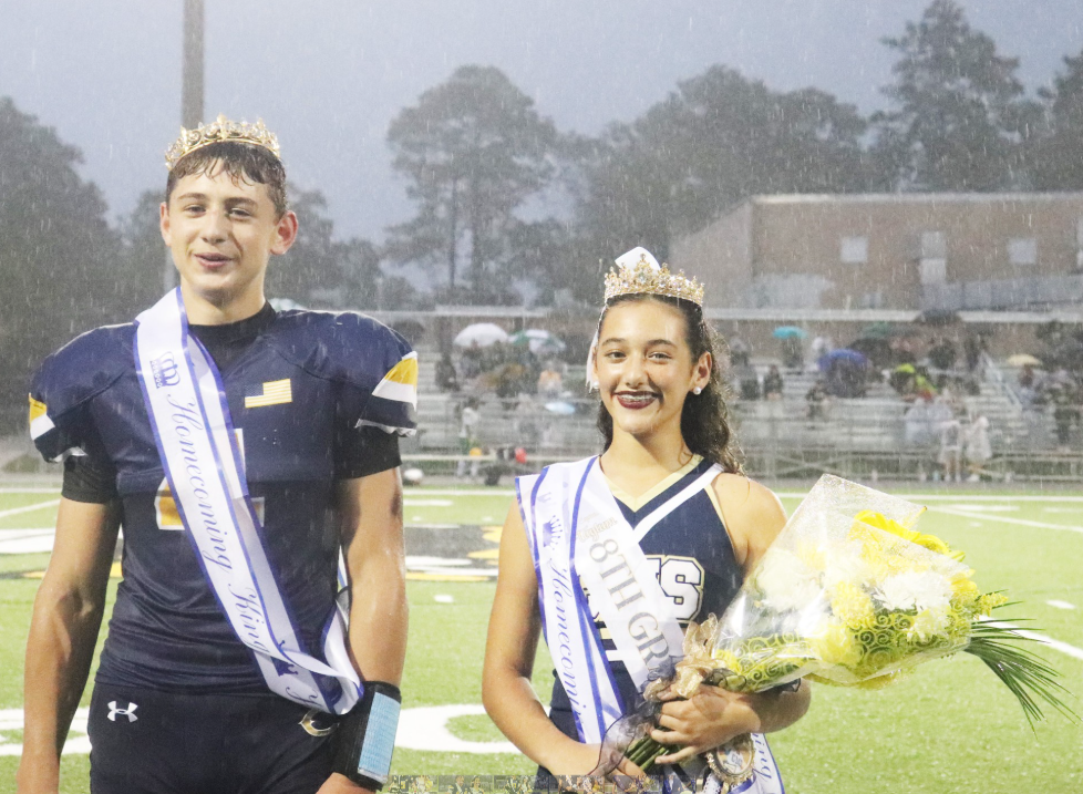 Two middle school homecoming representatives stand on a football field during a rainy evening ceremony. Both wear crowns and homecoming sashes. One student is dressed in a football uniform, while the other wears a cheer uniform and holds a bouquet of yellow and white flowers. Spectators with umbrellas are visible in the bleachers behind them, and stadium lights illuminate the wet field and falling rain.
