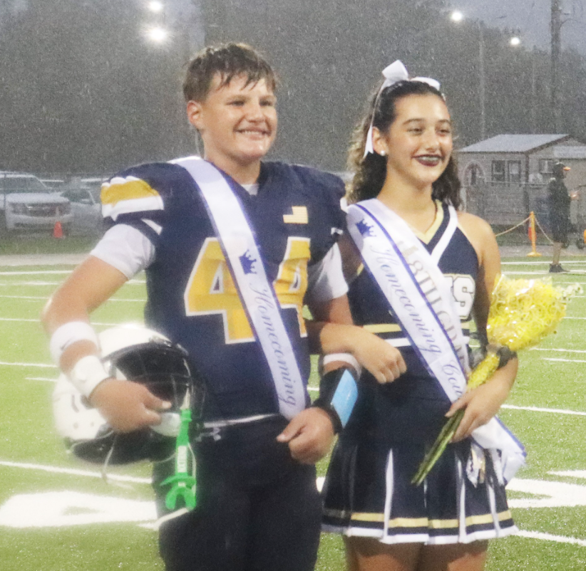 Two middle school homecoming court representatives stand together on a football field during a rainy evening event. One student wears a football uniform with a homecoming sash and holds a football helmet. The other student wears a cheer uniform with a homecoming court sash, carries a bouquet of yellow flowers, and has a white bow in their hair. Stadium lights illuminate the wet field, with vehicles and a small building visible in the background.