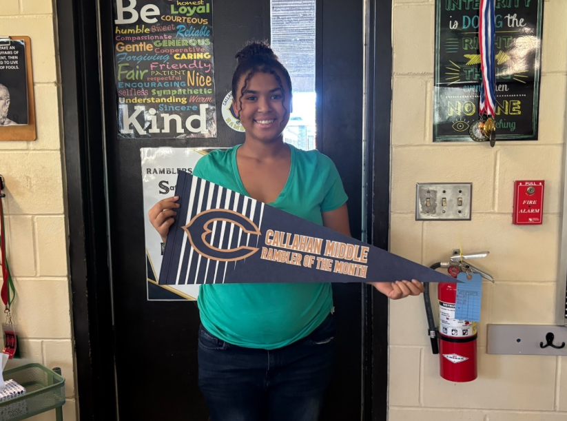 A student stands indoors holding a blue-and-white pennant that reads “Callahan Middle Rambler of the Month.” Behind the student is a classroom door decorated with character education posters, including one that features the words “Be Kind” surrounded by positive traits. Additional posters, medals, a fire alarm, and a fire extinguisher are visible on the wall nearby.
