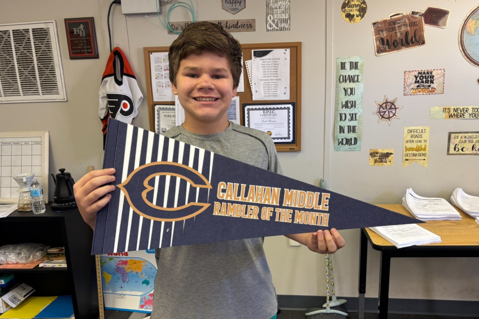 A student stands in a classroom holding a blue-and-white pennant that reads “Callahan Middle Rambler of the Month.” The classroom wall behind the student displays educational and motivational posters, certificates, and a bulletin board. A desk with stacks of papers, a world map, and classroom supplies are visible in the background.