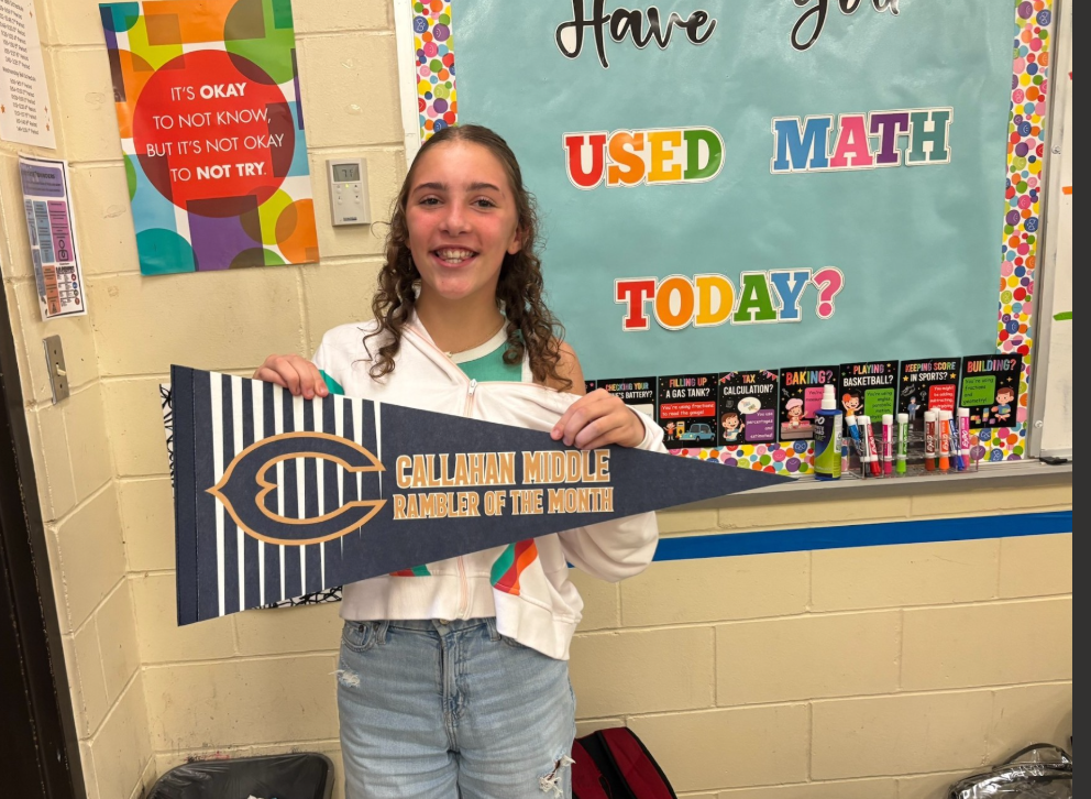 A student stands in a classroom holding a blue and white pennant that reads “Callahan Middle Rambler of the Month.” Behind the student is a bulletin board displaying the message “Have You Used Math Today?” along with math-themed decorations and classroom supplies. A poster on the wall reads, “It’s okay to not know, but it’s not okay to not try.”