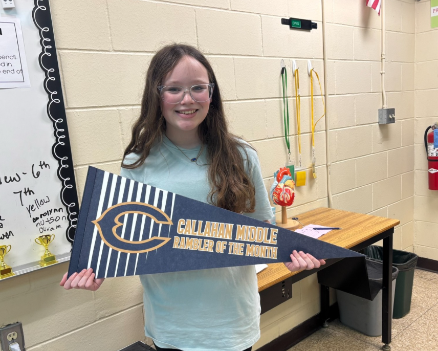 A student stands in a classroom holding a blue-and-white pennant that reads “Callahan Middle Rambler of the Month.” The classroom includes a whiteboard with handwritten notes and award trophies, a table displaying an anatomical heart model, and several lanyards hanging on the wall. A fire extinguisher is mounted on the wall in the background.
