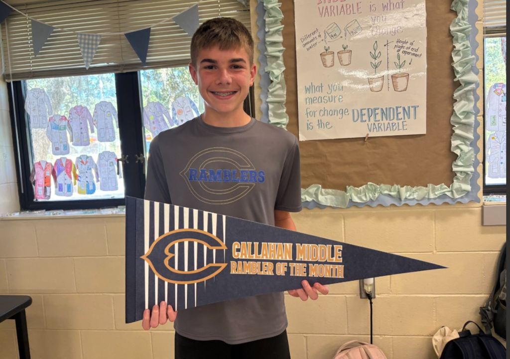 A student stands in a classroom holding a blue-and-white pennant that reads “Callahan Middle Rambler of the Month.” Behind the student is a science classroom bulletin board featuring a display about independent and dependent variables with plant growth illustrations. Windows decorated with student artwork and blue-and-white pennant banners are visible in the background. Provide your feedback on BizChat