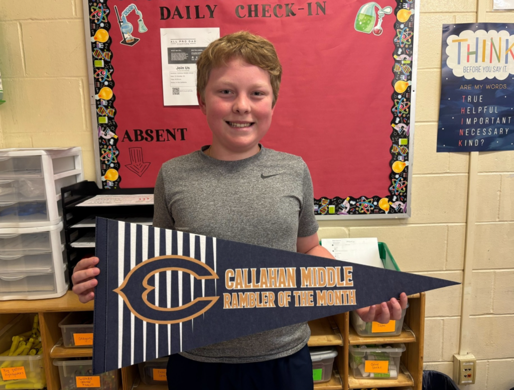 A student stands in a classroom holding a blue-and-white pennant that reads “Callahan Middle Rambler of the Month.” Behind the student is a red bulletin board labeled “Daily Check-In” with science-themed decorations and an “Absent” section. Classroom storage bins and shelves are visible in the background, along with a poster that reads, “Think before you say it. Are my words true, helpful, important, necessary, kind?”