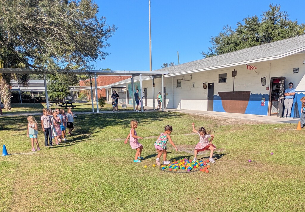 Children playing on a sunny school playground on a grassy lawn, with a colorful plastic balls in the middle of a red hula hoop on the ground and a white building and brick walkway in the background.