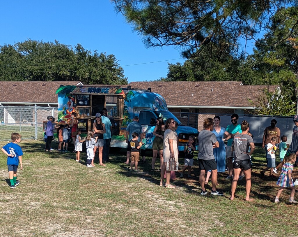 Kids and adults stand on a sunny school playground near a colorful ice-cream truck with an open serving window, with a few children walking toward the truck under pine trees.
