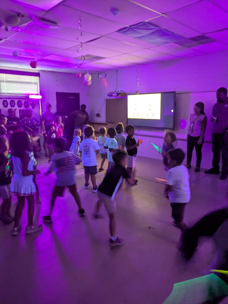 Children and a few adults dance and move around in a community room under purple stage lights, with a projector screen showing a bright image and balloons hanging from the ceiling.