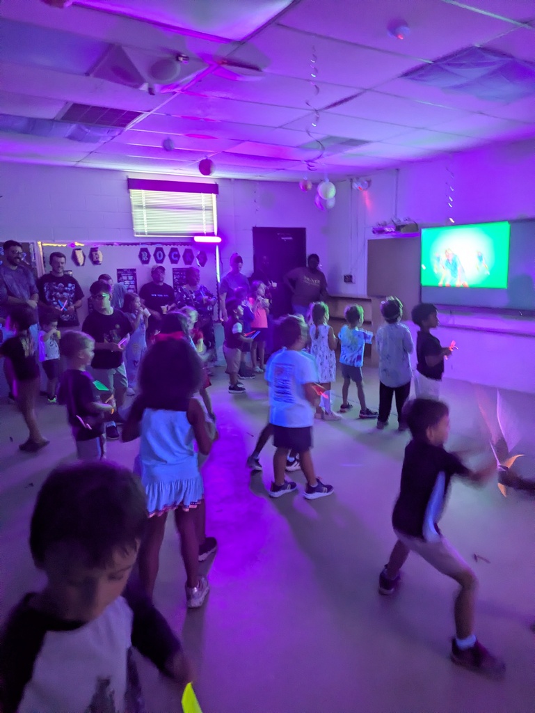 A group of children dance in a brightly lit indoor party room as a projector screen glows and colorful purple stage lights wash over the ceiling and walls.