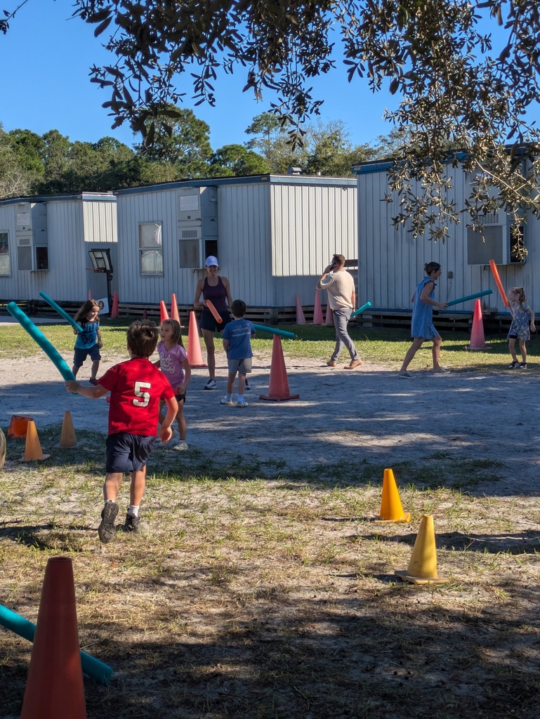 Children play outdoors in a sunny yard, running between bright orange and blue training cones near a blue metal building, while an adult in the background supervises.
