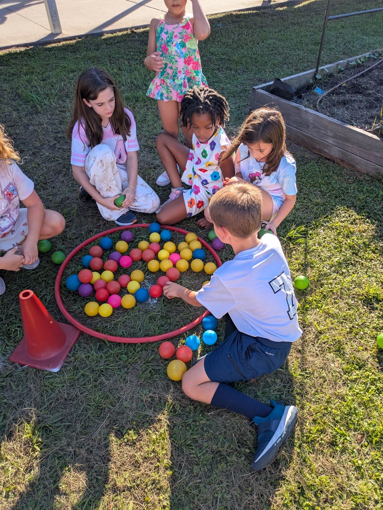 Several children in colorful outfits play outdoors on grass around a red ring filled with multicolored plastic balls, with a small watering can nearby and a fenced play area in the background.