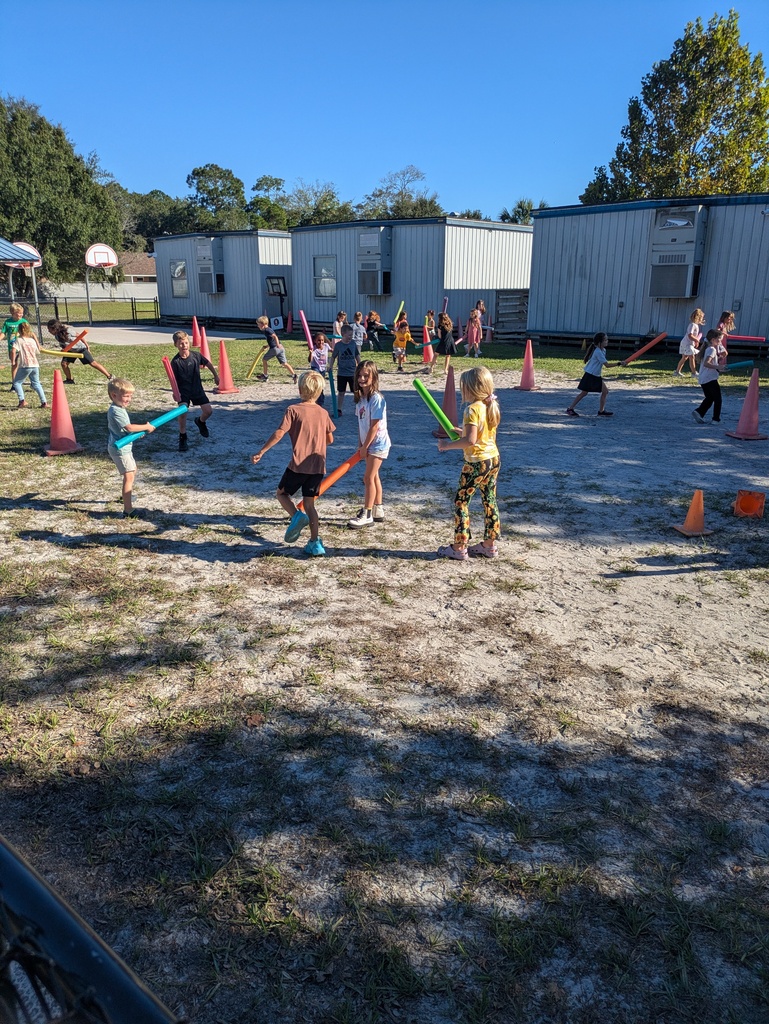 Children play in an outdoor activity area on a sunny day, moving through a playground course with colorful cones and pool noodles with blue storage buildings and trees in the background.