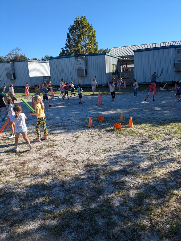 Children play outdoors in a sunny schoolyard, running and jumping around colorful cones and foam sticks while a blue metal building and trees sit in the background.