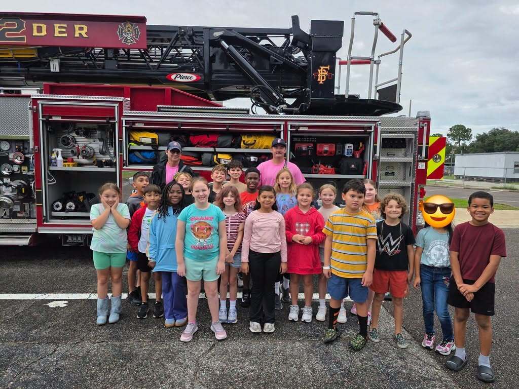 A large group of Emma Love Hardee Elementary School students and firefighters pose in front of an open fire truck displaying hoses, equipment, and emergency gear.