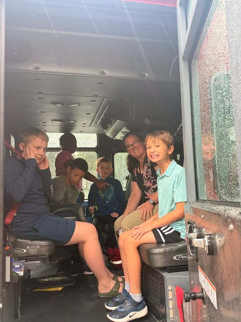Students and a staff member sit inside the cab of a fire truck while learning about firefighting equipment and emergency response.