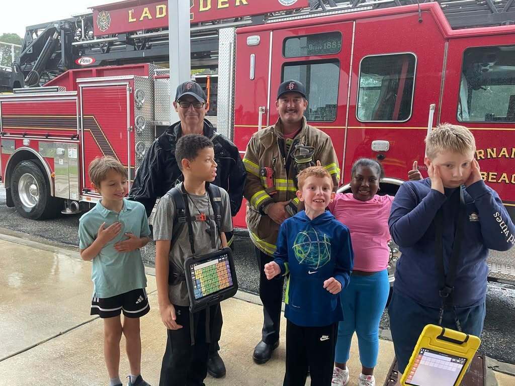 Elementary students stand with two firefighters in front of a red ladder truck during a visit from the Fernandina Beach Fire Department.