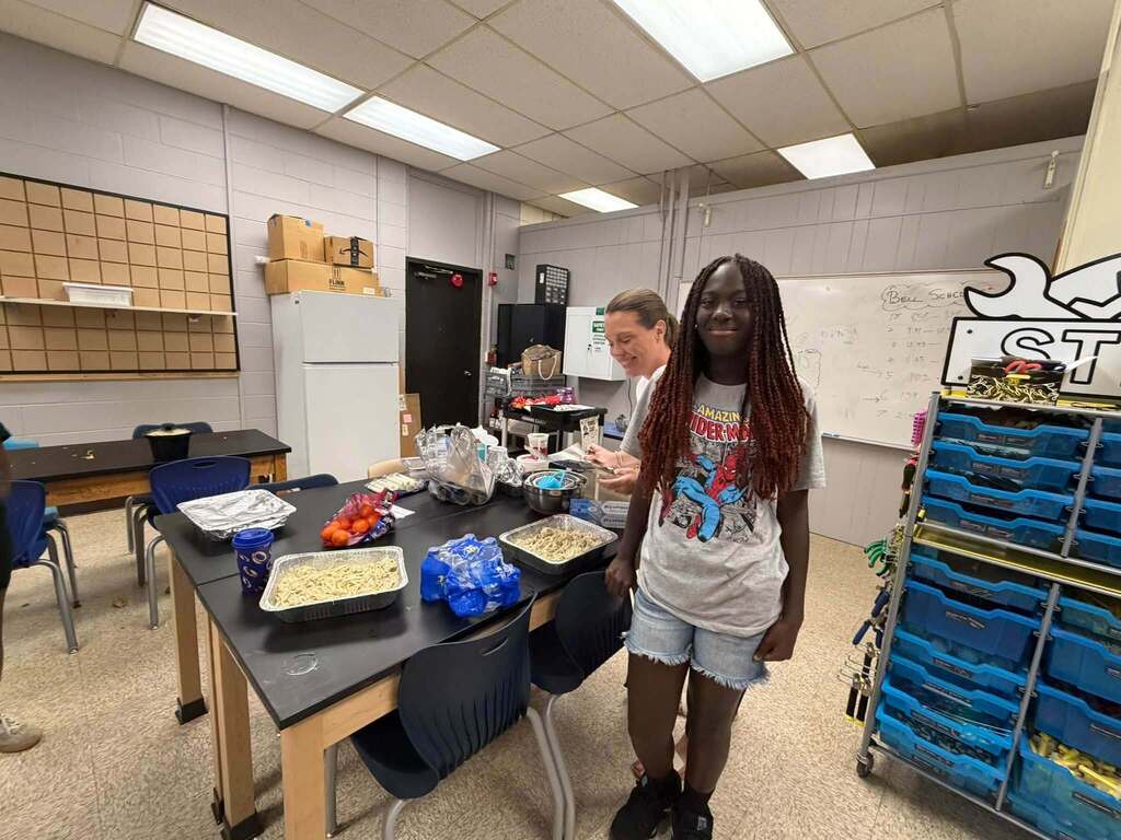 Student stands beside a classroom worktable filled with ingredients, pans, bottled water, and cooking supplies while an adult helps prepare food during Open Kitchen Club.