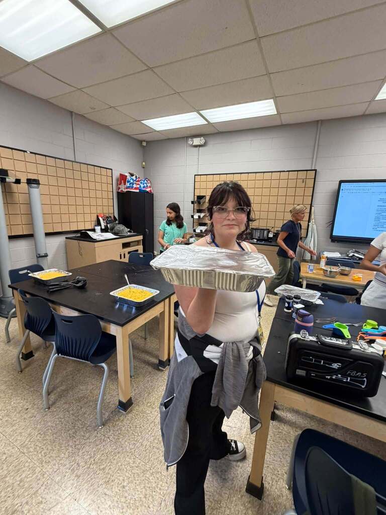 Student holds a covered foil pan in the Fernandina Beach Middle School STEM lab, with additional prepared dishes and cooking supplies visible on classroom tables.