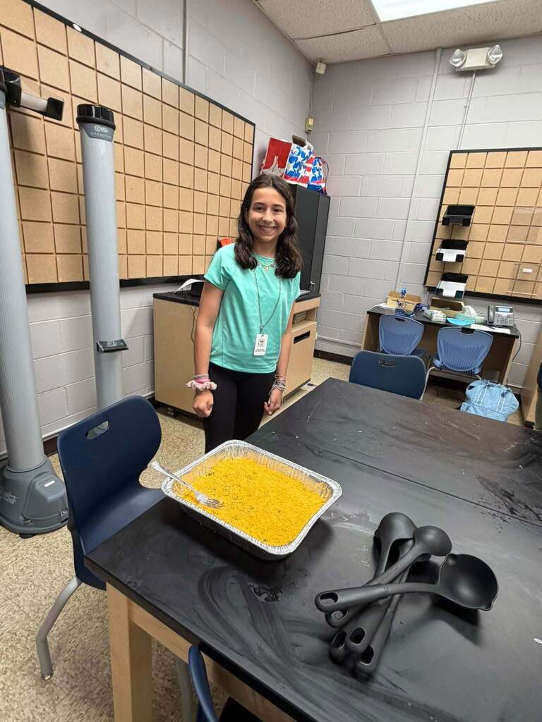 Student stands beside a prepared pan of food and cooking utensils on a STEM lab table during a weekly Open Kitchen Club activity.