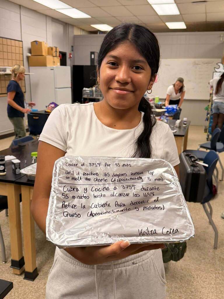Student smiles while holding a covered meal pan labeled with cooking instructions written in English and Spanish during Fernandina Beach Middle School’s Open Kitchen Club.