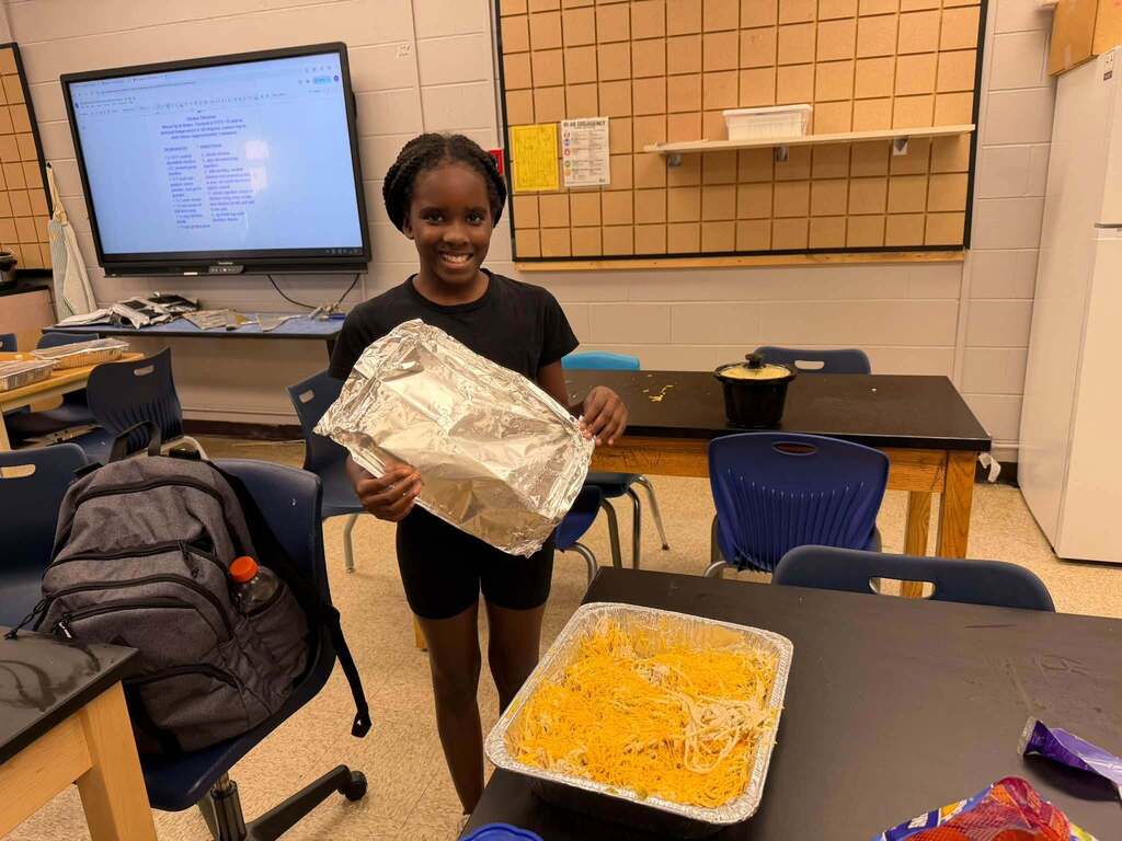 Fernandina Beach Middle School student smiles while holding a foil pan cover beside a large pan of a prepared pasta dish in the school STEM lab during Open Kitchen Club.