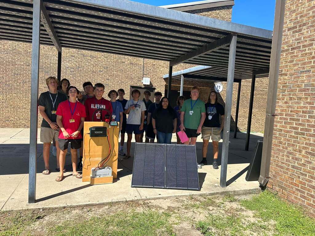 Fernandina Beach High School Electricity students stand outdoors behind a portable solar generator they built, with a large solar panel, battery, inverter, wiring, and mobile equipment assembly displayed in front of the group.