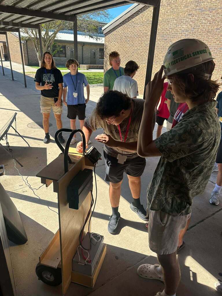 Fernandina Beach High School Electricity students gather around their portable solar generator outdoors while examining its electrical components. One student wears a hard hat as classmates observe the battery, wiring, inverter, and other equipment used in the hands-on solar energy project.
