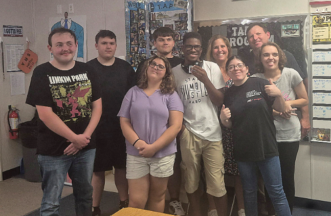 A group of students and staff members pose together in a classroom, smiling at the camera. Classroom posters, bulletin boards, and instructional materials are visible in the background.