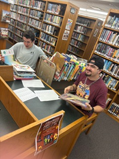 Two Hilliard Middle-Senior High School students work at a table inside the Hilliard Branch Library during a Work-Based Learning Experience. Both students are seated among library bookshelves while organizing and reviewing books and work materials.