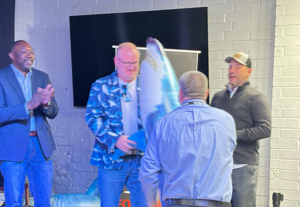 On an indoor stage, an individual in a blue shark-patterned jacket presents a large plush shark to another individual standing with their back to the camera. Two additional individuals stand nearby, one applauding and another observing. Shark-themed decorations are visible on and around the stage, including another plush shark near the floor. The stage is set against a light-colored brick wall with a large dark display screen in the background. Bright stage lighting illuminates the presentation.