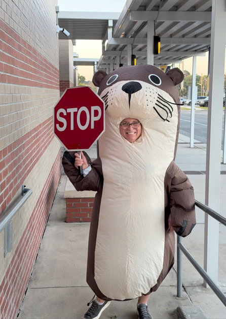 A smiling school staff member wearing an inflatable brown otter costume holds a red STOP sign outside the school near the student drop-off area.