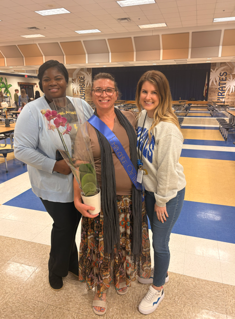 Three smiling Southside Elementary School staff members pose together in the cafeteria. The staff member in the center wears an Employee of the Month sash and holds a potted purple orchid as her colleagues celebrate her recognition.