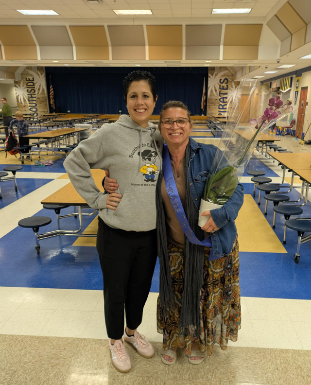 Two smiling Southside Elementary School staff members stand together in the cafeteria. One wears a Southside Elementary sweatshirt, while the other wears an Employee of the Month sash and holds a potted purple orchid in celebration of her recognition.