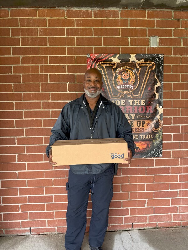 School Resource Officer Caldwell stands in front of a brick wall holding a Food for Good by PepsiCo box while preparing to deliver food to a family.