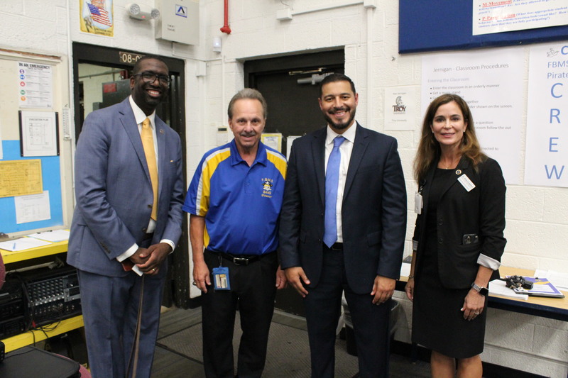 The Commissioner of Education and district leaders pose with a teacher in a middle school band classroom, standing in front of posters and classroom procedures displayed on the wall.