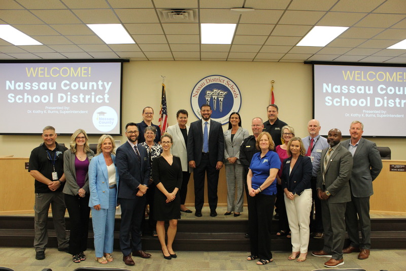 Group photo of the Florida Commissioner of Education standing with Nassau County School District leadership inside the district office boardroom. Two large screens behind the group display a welcome message for the Commissioner.