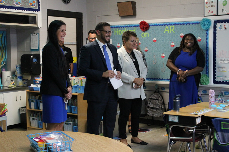 The Commissioner of Education, Superintendent Dr. Kathy Burns, and other district leaders stand inside an elementary classroom, smiling and engaging with staff.