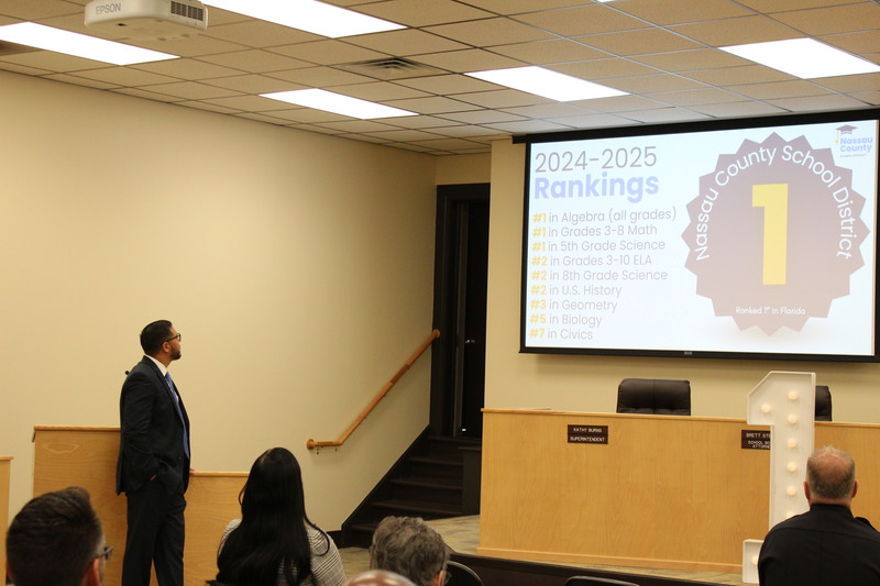 The Florida Commissioner of Education stands at the front of the Nassau County School District boardroom, looking at a projected slide that highlights the district’s 2024–2025 rankings, including its #1 position in Florida.