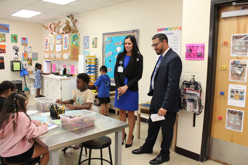 The Commissioner of Education and district staff visit an elementary art classroom, speaking with students seated at tables working on drawing activities.