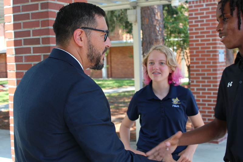 The Commissioner of Education greets two middle school students outdoors on campus, shaking hands with one student while another looks on.