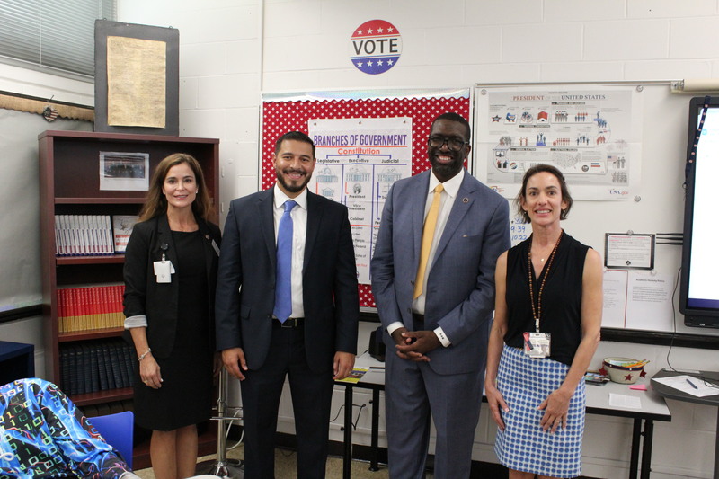 The Commissioner of Education and district officials pose for a photo with a teacher inside a middle school social studies classroom. A poster on the wall shows the branches of government.