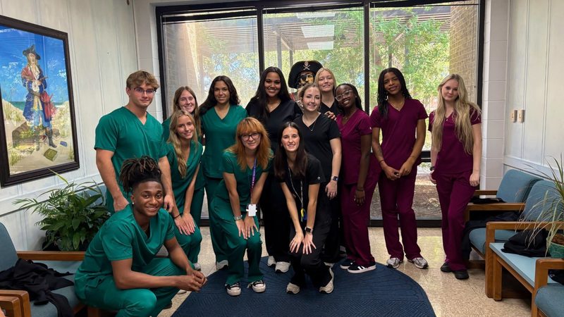 CNA students in green, black, and maroon scrubs pose together indoors, representing a healthcare training program.