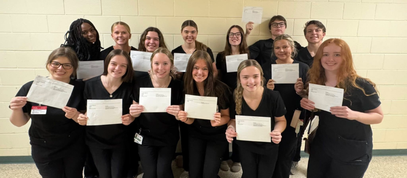 CNA students in black scrubs hold documents and pose together in a classroom after completing certification requirements.