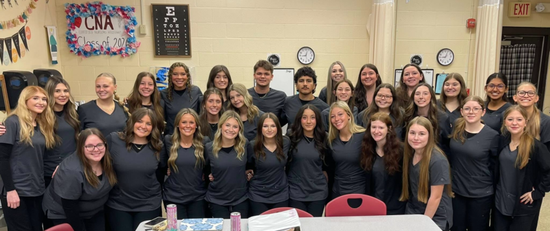 Group of CNA students in black scrubs pose together in a classroom with medical training equipment and a “CNA Class” sign on the wall.